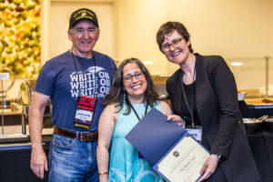 SFWA CFO Jonathan P. Brazee (left) and SFWA President (Kate Ristau) celebrate Nebula Finalist Jennifer Hudak. (Photo Credit: Kaitrin Acuna, 2025.)